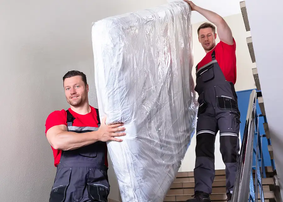 Two Moscardelli movers in red uniforms carefully carrying a plastic-wrapped mattress down a staircase of an apartment building in Springfield, IL