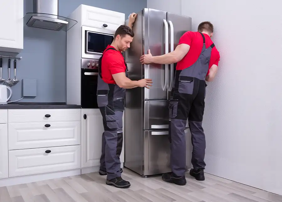 Two Moscardelli Moving & Storage movers in red uniforms carefully repositioning a stainless steel refrigerator in a white kitchen in Springfield, IL