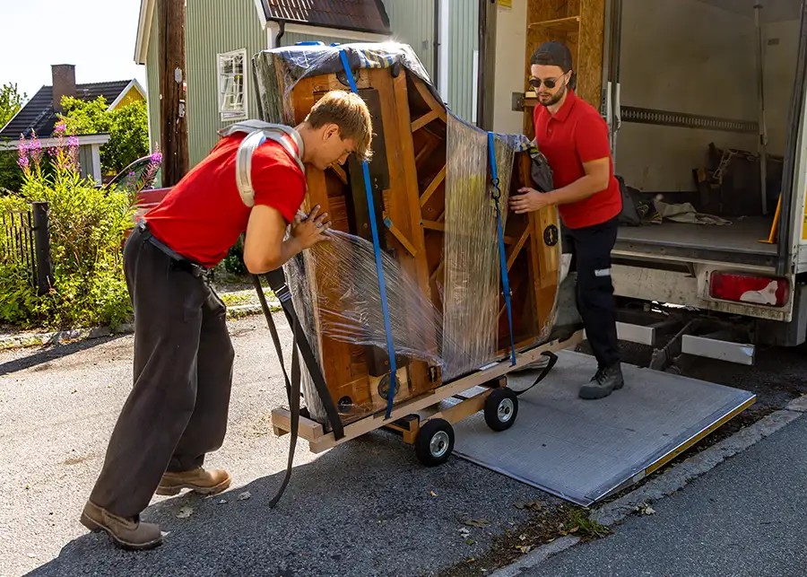 Two Moscardelli Moving & Storage movers in red shirts using straps and a flatbed dolly to load a wrapped baby grand piano onto a moving truck in Springfield, IL