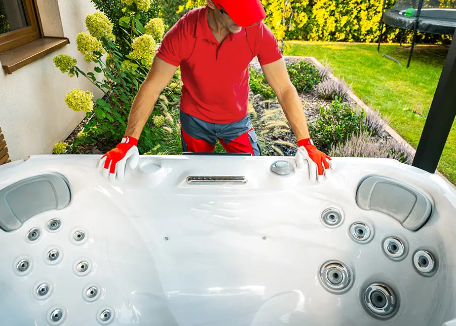 A Moscardelli mover in a red uniform and gloves carefully maneuvering a large white hot tub across a landscaped backyard in Springfield, IL