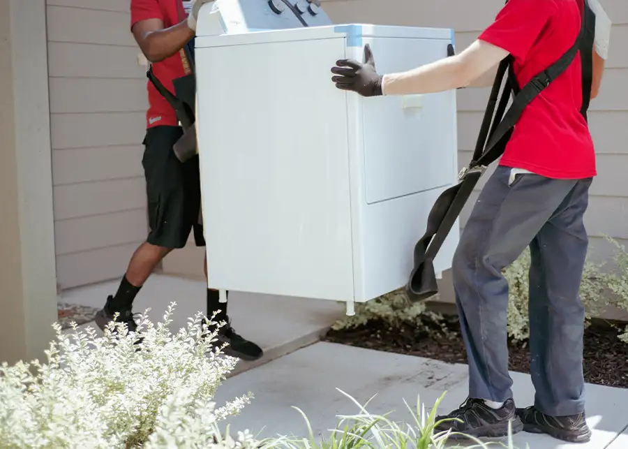 Two Moscardelli Moving & Storage movers using lifting straps to carry a top-load washing machine appliance along a residential walkway in Springfield, IL